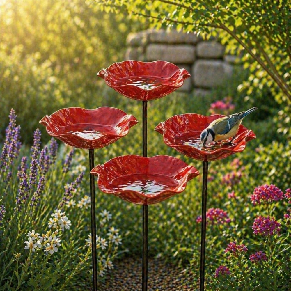 Rood vogelbad in bloemvorm met golvende rand op een metalen steel tussen bloeiende tuinplanten